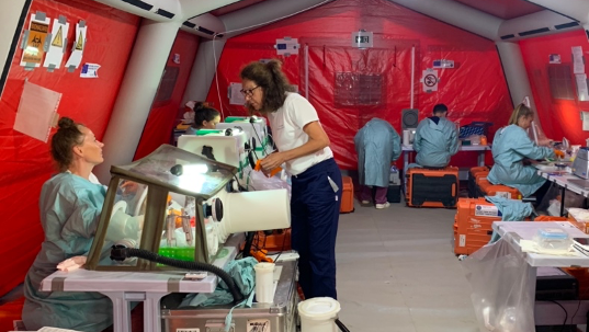 EMLab team members worked inside a field laboratory setup inside an inflatable tent during the EU MODEX. The image shows a laboratory in a red tent with staff in lab coats working on benches and with glove boxes.