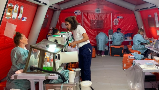 The image shows a laboratory in a red tent with staff in lab coats working on benches and with glove boxes.