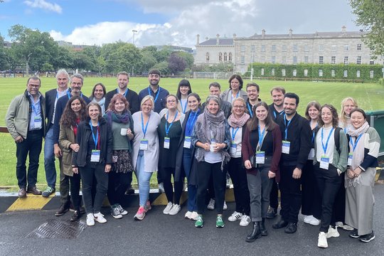 Members of the Sex Differences in Immunity research group stand outside a grassy area and Trinity College in Dublin. They all look cheerful and wear a conference badge around their necks