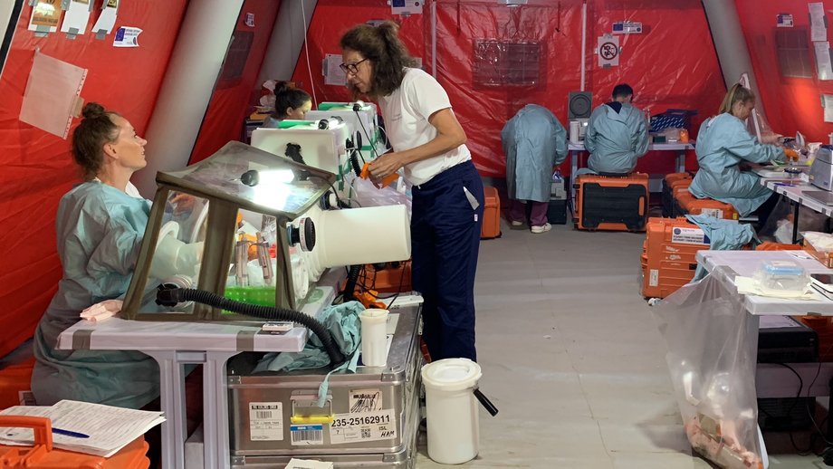 Das Foto zeigt ein Labor in einem roten Zelt mit Mitarbeitern in Laborkitteln, die an Bänken und mit Handschuhschachteln arbeiten.
