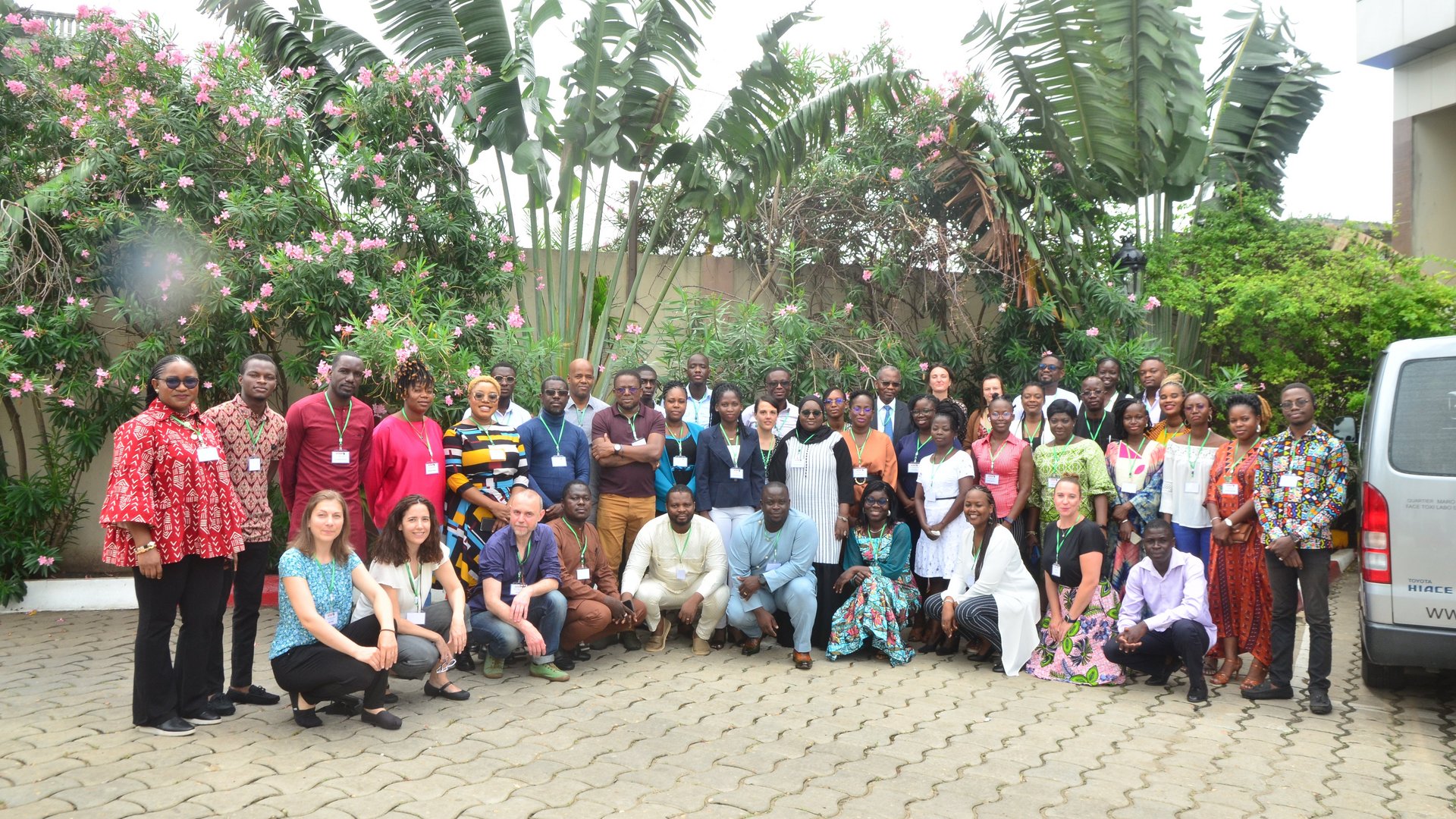 Group picture of ASAAP team at annual meeting. Gruppenbild des ASAAP-Teams bei der Jahrestagung: zu sehen ist eine große diverse Gruppe vor einem Hintergrund mit Palmen und bunten Blumen. Die Gruppe schaut in Richtung der Kamera und ist in insgesamt drei Reihen aufgestellt.