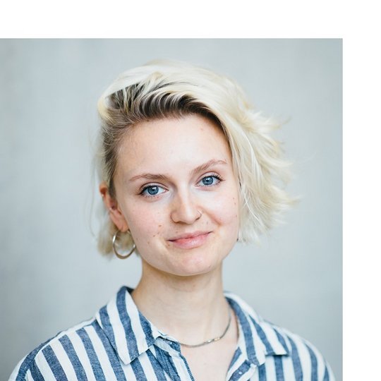 Portrait of a white young person with a blonde bob wearing a striped shirt - smiling into the camera and standing against a concrete wall.