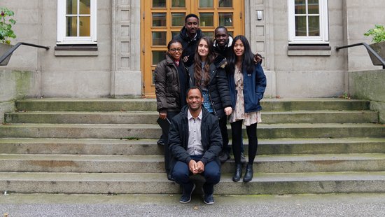 Six people wearing winter clothing stand and crouch on stone steps in front of a building with two windows and a wooden door.