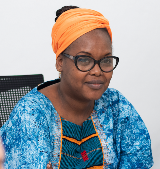 Porträtfoto der erfahrenen Forscherin Dr. Oumou Maïga-Ascofaré. Sie trägt ein oranges Kopftuch, eine dunkel umrandete Brille, ein dunkelblaues Oberteil und eine gemusterte hellblaue Jacke.
