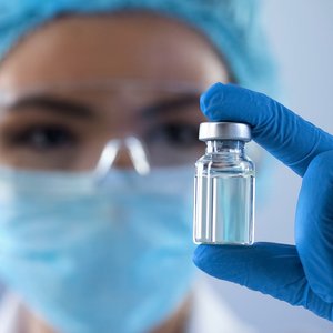 Foto einer Person mit Mund-Nasenschutz und blauen Laborhandschuhen und einer Impfampulle in der Hand