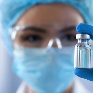 A photo of a person wearing a face mask and blue laboratory gloves, holding a vial of vaccine
