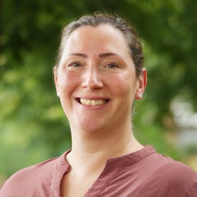 Dr Christine Jacobsen: a woman with dark hair tied back. She wears a brown-red shirt and stands in front of a blurred leafy backgrou