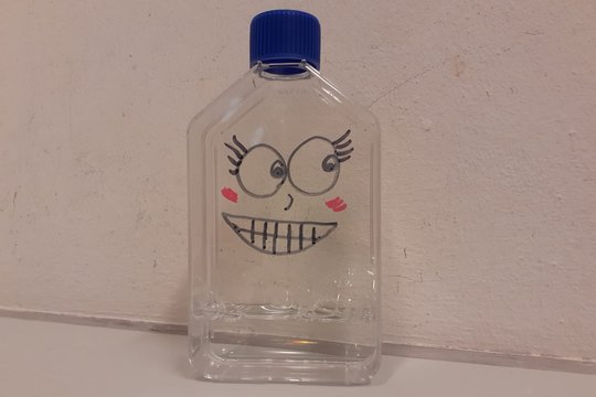 Monja Paasche Eine durchsichtige, eckige Zellkulturflasche mit blauem Deckel. Auf die Zellkulturflasche sind große Augen mit Wimpern, eine kleine Nase, rote Wangen und ein lachender Mund mit Zähnen gemalt.