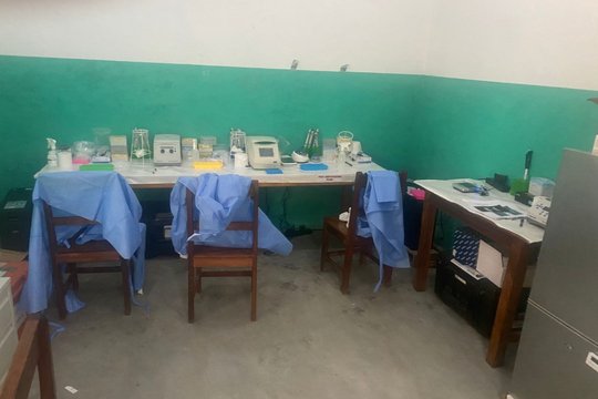 Interior of the new sequencing laboratory at LFHV-GKD. Guéckédou, Guinea, December 2025. One big table with many different laboratory equipment and three chairs in front of it. On the right side is another table with the sequencing device on it and a fridge.