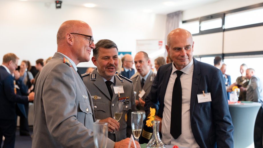 Stehempfang Das Foto zeigt im Vordergrund drei Herren an einem Stehtisch. Zwei tragen Uniformen, einer einen Anzug. Im Hintergrund sind weitere Personen in einem hellem Raum zu sehen.