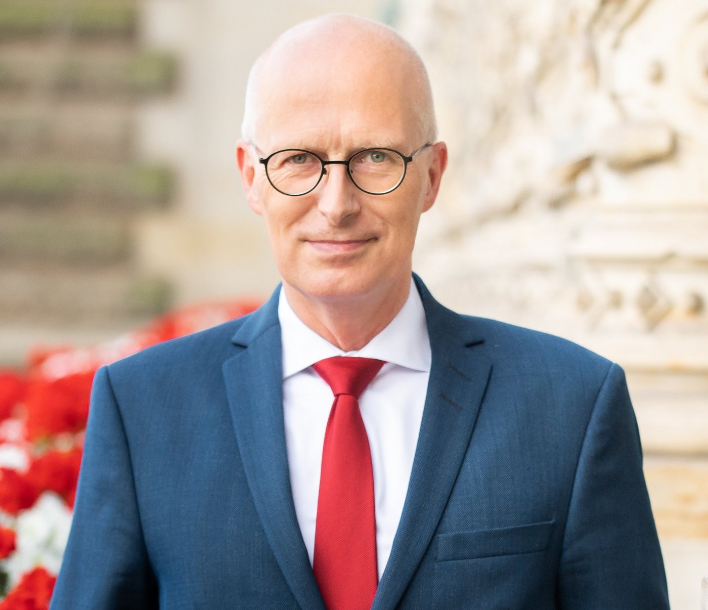 Ein Mann in einem blauen Anzug mit roter Krawatte steht selbstbewusst vor einem historischen Gebäude, das mit kunstvollen Schnitzereien geschmückt ist. In der Nähe blühen leuchtend rote Blumen.