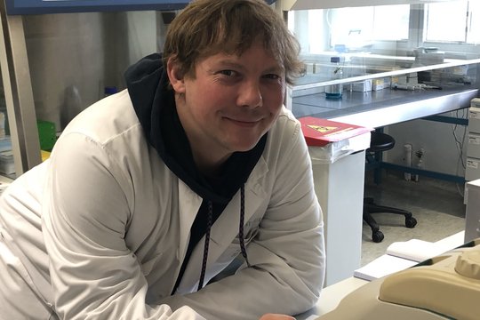 Philipp Böhmer Philipp Böhmer: in Forscher im Labor lächelt in die Kamera. Er steht auf einem Tisch aufgelehnt, trägt einen weißen Kittel, darunter einen dunkelblauen Kaputzenhoodie, sowie halblanges blondes Haar.