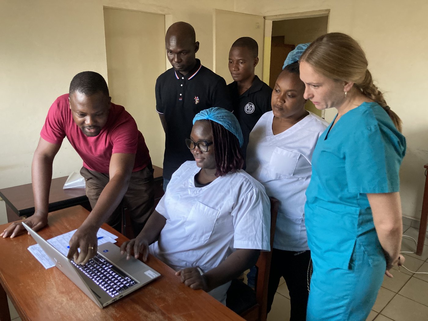 Analysing LASV RT-PCR runs and reporting results A lab group of five local staff from Benin plus one German trainer are looking at a laptop, one of them is pointing to the screen, highlighting something.