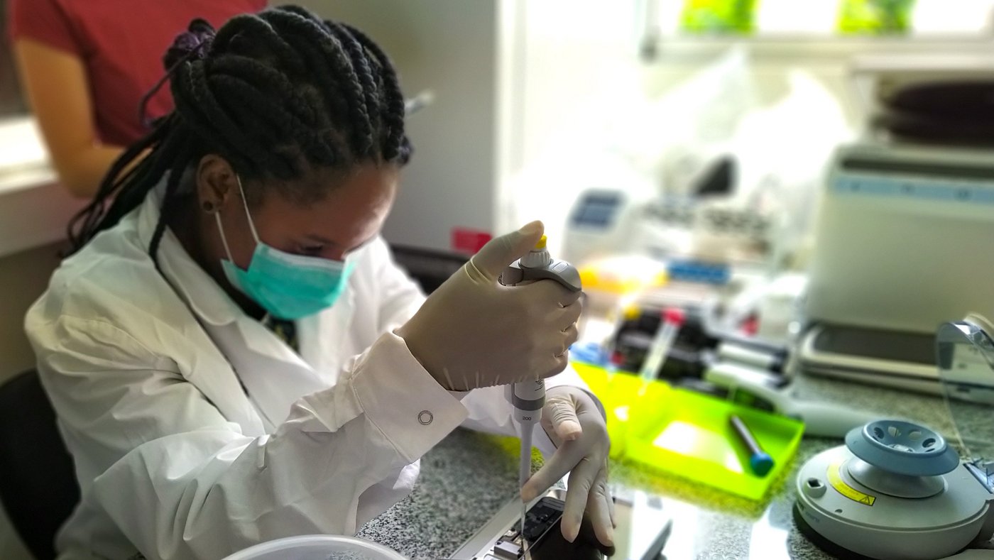 Das Foto zeigt eine Laborexpertin, die an einem Labortisch sitzt. Sie pipettiert eine Flüssigkeit mit einer Mikropipette in ein Sequenziergerät.