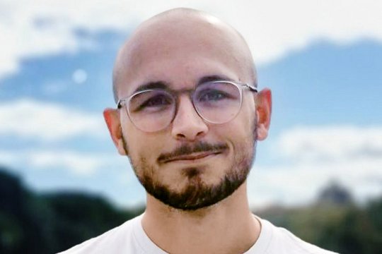 Louis Sarrazin Louis Sarrazin: Ein Doktorand steht lacht freundlich in die Kamera. Er trägt eine Glatze, einen schwarzen Kinn- und Oberlippenbart, ein weißes Shirt und eine runde Brille. Er steht draußen vor einem blauen, aber leicht wolkigem Himmel.