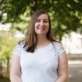 Glaucia Herzer: eine lächelnde Frau mit langen mittelblonden Haaren. Sie trägt ein weißes T-Shirt und steht auf einem sonnigen Weg mit Bäumen.