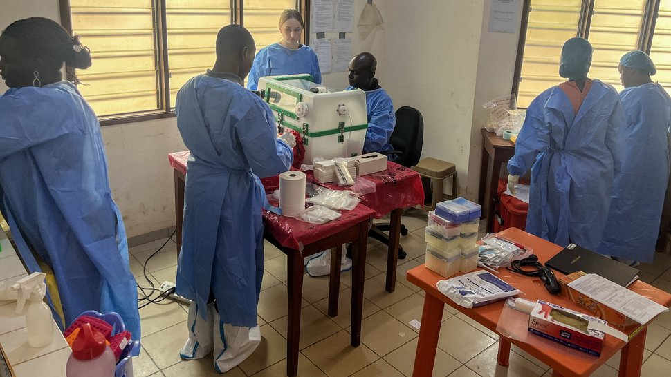 Foto mehrerer internationaler Wissenschaftler:innen in einem Raum mit Lamellenfenstern. Zwei Kollegen arbeiten an der Glove box.