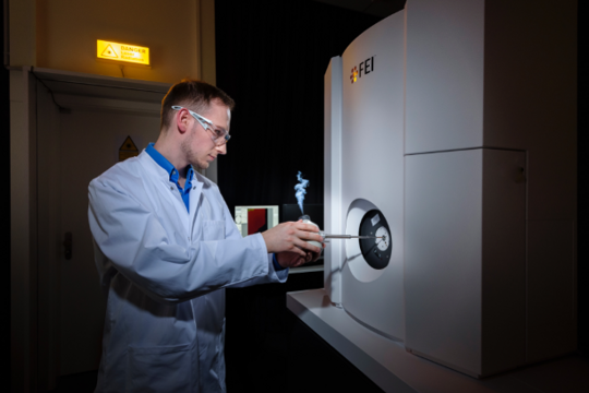 Krankheitserreger erforschen Eine Person in Laborkittel und Schutzbrille lädt in einer Laborumgebung eine Probe in ein großes wissenschaftliches Instrument mit der Aufschrift FEI.