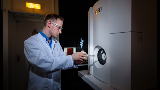 A person in a lab coat and safety glasses loads a sample into a large scientific instrument labeled FEI in a laboratory setting.