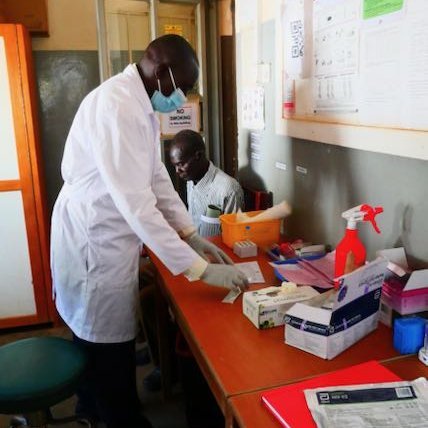 Covid 19 Testing at Bobbi Hospital, Uganda Eine Person steht an einem Tisch, auf dem Laborutensilien liegen, und arbeitet. Er trägt Handschuhe, eine Nasen-Mund-Bedeckung und einen Laborkittel. Eine weiter Person sitzt auf einem Stuhl neben dem Tisch.