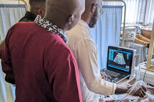 The picture shows three men standing over a volunteer who is being scanned with an ultrasound device.