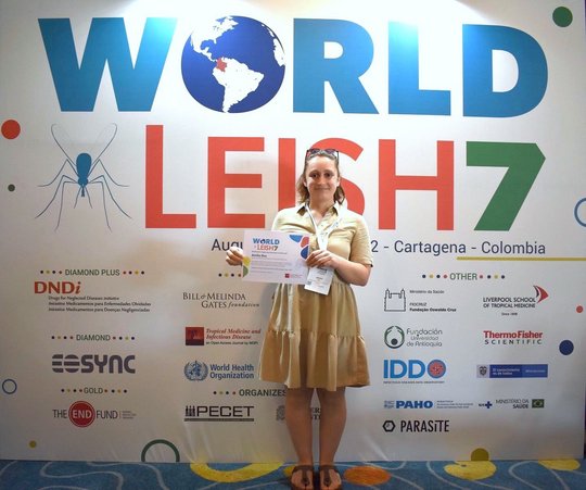 Eine Forscherin hält ein Urkunde in den Händen. Sie trägt ein knielanges, beigefarbenes Sommerkleid und eine in die Haare gesteckte Sonnenbrille. Hinter ihr ist ein großer Schriftzug "WorldLeish7" zu sehen, darunter mehrere Logos von verschiedenen Kooperationspartnern.