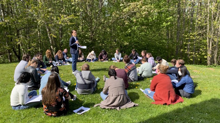 Eine Gruppe internationaler Teilnehmender eines Workshops sitzt in einem Kreis auf einer Wiese, in der Mitte steht eine Kursleiterin und erläutert Inhalte des Workshops.
