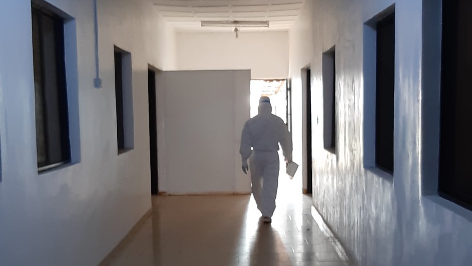 Das Foto zeigt einen weißen, leeren Flur, mit Fenstern an den Seiten zu Behandlungsräumen. Am Ende des Gangs kommt eine Person in einem weißen Ganzkörperschutzanzug auf einen zu.