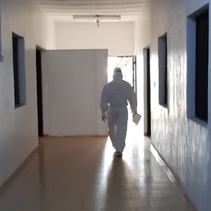 Das Foto zeigt einen weißen, leeren Flur, mit Fenstern an den Seiten zu Behandlungsräumen. Am Ende des Gangs kommt eine Person in einem weißen Ganzkörperschutzanzug auf einen zu.