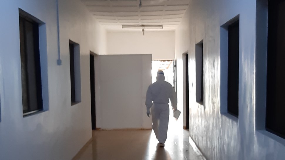 Das Foto zeigt einen weißen, leeren Flur, mit Fenstern an den Seiten zu Behandlungsräumen. Am Ende des Gangs kommt eine Person in einem weißen Ganzkörperschutzanzug auf einen zu.