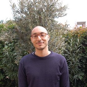 A man with a shaved head and glasses wearing a dark sweater, smiling while standing in front of a lush green backdrop of foliage.