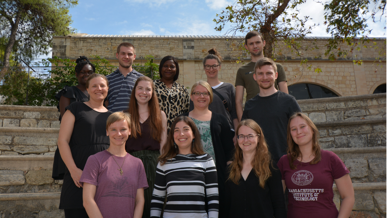 Gruppenfoto der Laborgruppe. Die Mitglieder der Arbeitsgruppe stehen lächelnd neben- und hintereinander zusammen. Im Hintergrund sind Bäume und ein Gebäude zu sehen.