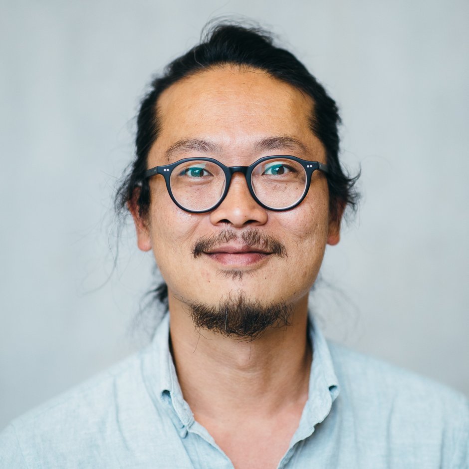 Portrait of a male person of color with glasses and dark hair pulled back, wearing a blue shirt.