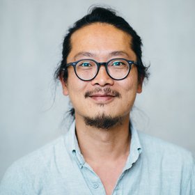 [Translate to English:] Portrait of a male person of color with glasses and dark hair pulled back, wearing a blue shirt.