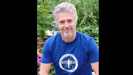 Ein Mann mit kurzen grauen Haaren und Bart sitzt im Freien. Er trägt ein blaues T-Shirt mit der Aufschrift „Tropeninstitut Hamburg“. Im Hintergrund sind grüne Pflanzen und rosa Blumen zu sehen.