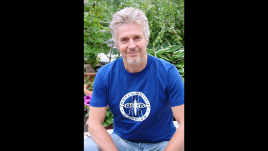 Ein Mann mit kurzen grauen Haaren und Bart sitzt im Freien. Er trägt ein blaues T-Shirt mit der Aufschrift „Tropeninstitut Hamburg“. Im Hintergrund sind grüne Pflanzen und rosa Blumen zu sehen.