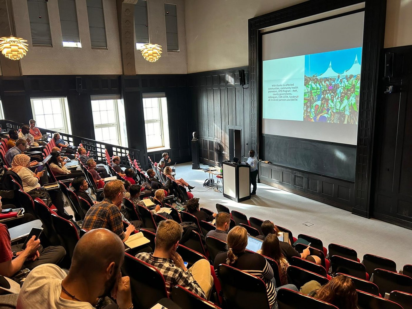 The picture shows a crowded lecture hall and a lecturer at the podium.