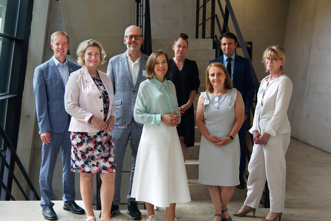 Photo of eight middle-aged men and women in a modern stairwell made of exposed concrete. They are dressed in summery clothes and look smilingly into the camera.