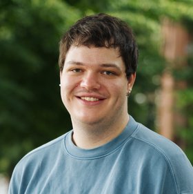 Robert Wildtraut: a man with dark, short hair in a blue sweatshirt smiles at the camera. In the background are blurred leaves.