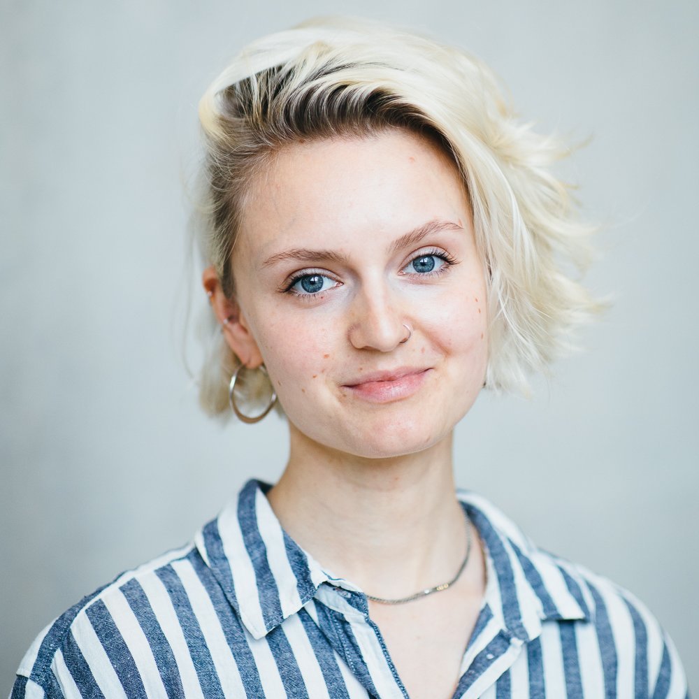 Portrait of a white young person with a blonde bob wearing a striped shirt - smiling into the camera and standing against a concrete wall.