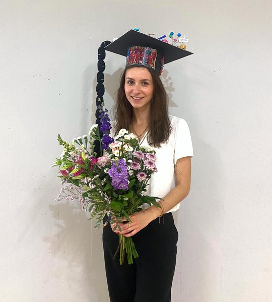 Melanie Lütkemeyer stands in front of a wall in the foyer of the Institute of Plant Sciences and Microbiology after her defence. She proudly wears her doctoral hat and carries a colourful bouquet of flowers