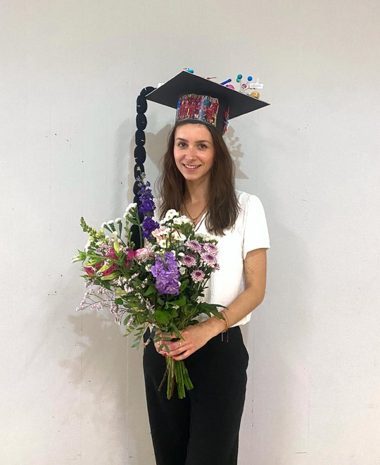 Melanie Lütkemeyer stands in front of a wall in the foyer of the Institute of Plant Sciences and Microbiology after her defence. She proudly wears her doctoral hat and carries a colourful bouquet of flowers