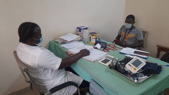 Zu sehen ist ein Arzt und ein Patient aus Burkina Faso, die sich gegenüber an einem Tisch mit unterschiedlichen Untersuchungsgeräten und Unterlagen sitzen.