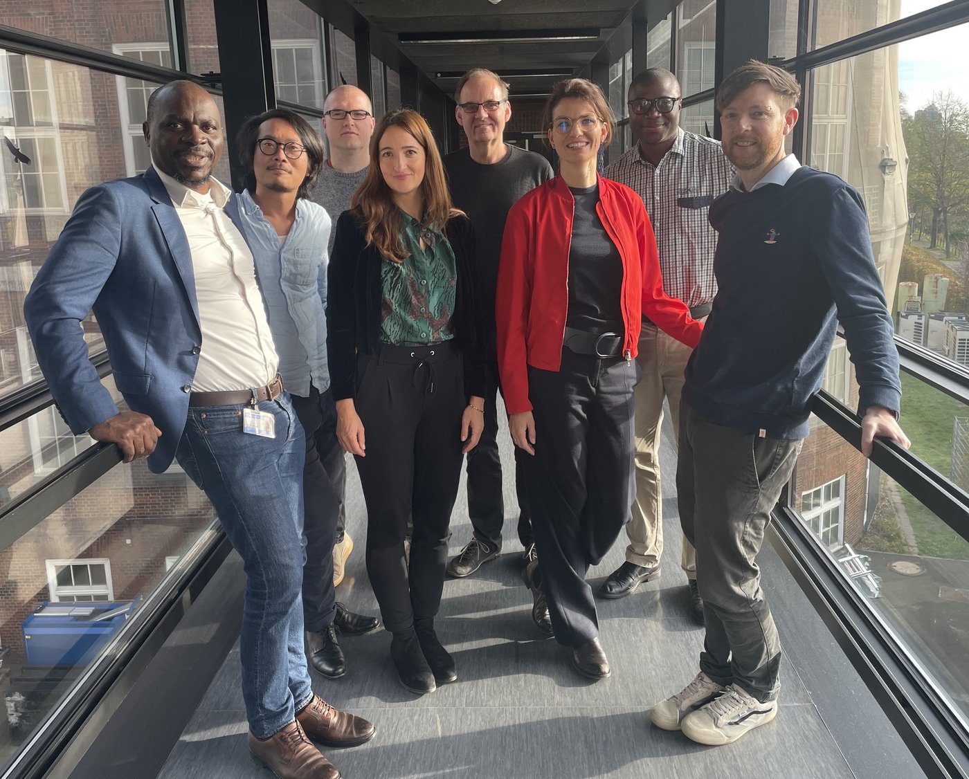 Six men and women with African and European looks are standing on the bridge between the two BNITM-buildings, smiling.