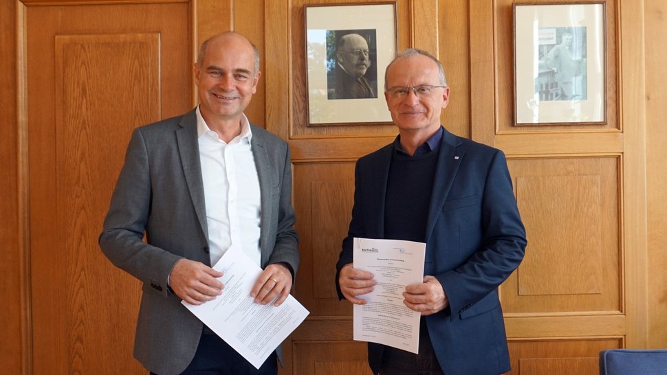 Prof. Jürgen May (li.) und Prof. Thomas Mettenleiter (re.) nach der Unterzeichnung des MoU Zwei freundliche Herren mittleren Alters, gekleidet in Hemd und Jackett, stehen vor einer mit hellem Holz vertäfelten Wand und lächeln freundlich. Beide halten den unterschriebenen MoU in Händen.