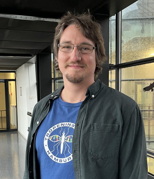 Ein Wissenschaftler mit kurzen braunen Haaren und einer Brille steht im Flur. Er trägt ein blaues T-Shirt mit einem Tropeninstitut-Logo, darüber ein offenes schwarzes Hemd.