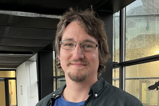 Gabor Endre Toth Ein Wissenschaftler mit kurzen braunen Haaren und einer Brille steht im Flur. Er trägt ein blaues T-Shirt mit einem Tropeninstitut-Logo, darüber ein offenes schwarzes Hemd.