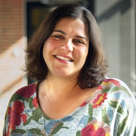 Dr Muna Affara The photo shows the researcher Dr Muna Affara. She wears half-length dark hair, a colourful jumper printed with large flowers and smiles friendly into the camera.