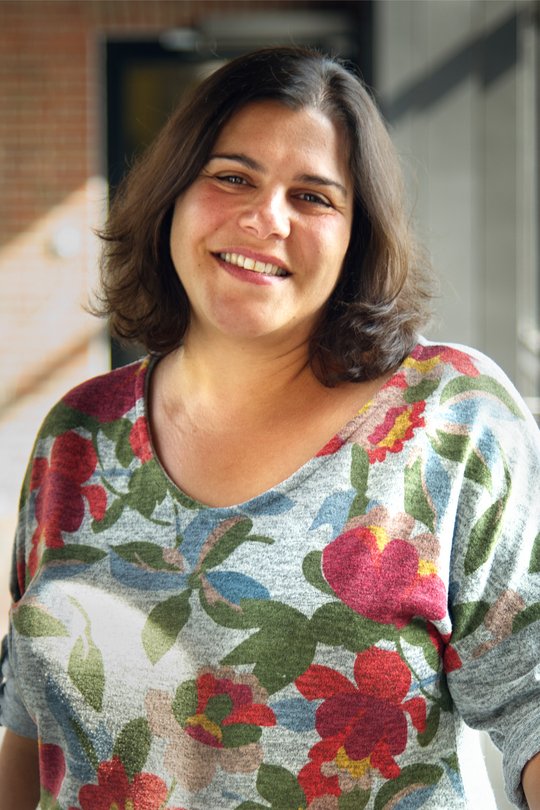 Dr. Muna Affara Das Foto zeigt die Forscherin Muna Affara. Sie trägt halblange dunkle Haare, einen bunten mit großen Blumen bedruckten Pullover und lächelt freundlich in die Kamera.