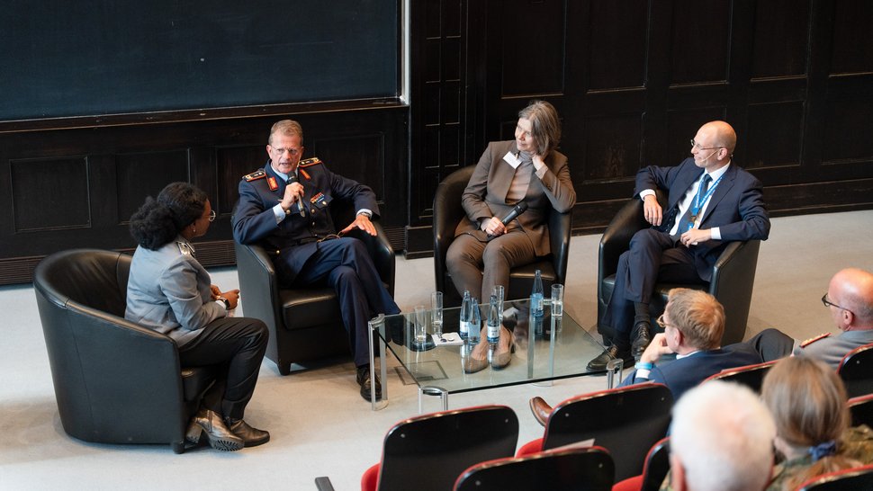 Podiumsdiskussion Das Foto zeigt vier Personen, die im Hörsaal auf vier dunklen Sesseln im Halbkreis sitzen.